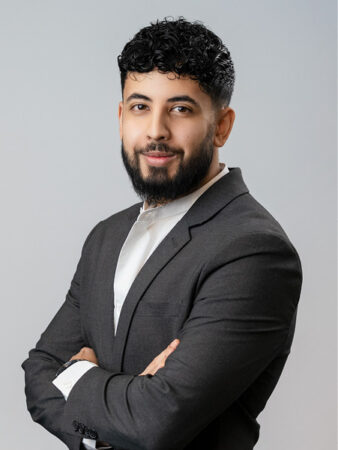Un homme aux cheveux courts bouclés et à la barbe, vêtu d'un costume sombre et d'une chemise blanche, se tient debout, les bras croisés, regardant l'appareil photo sur un fond clair et uni.