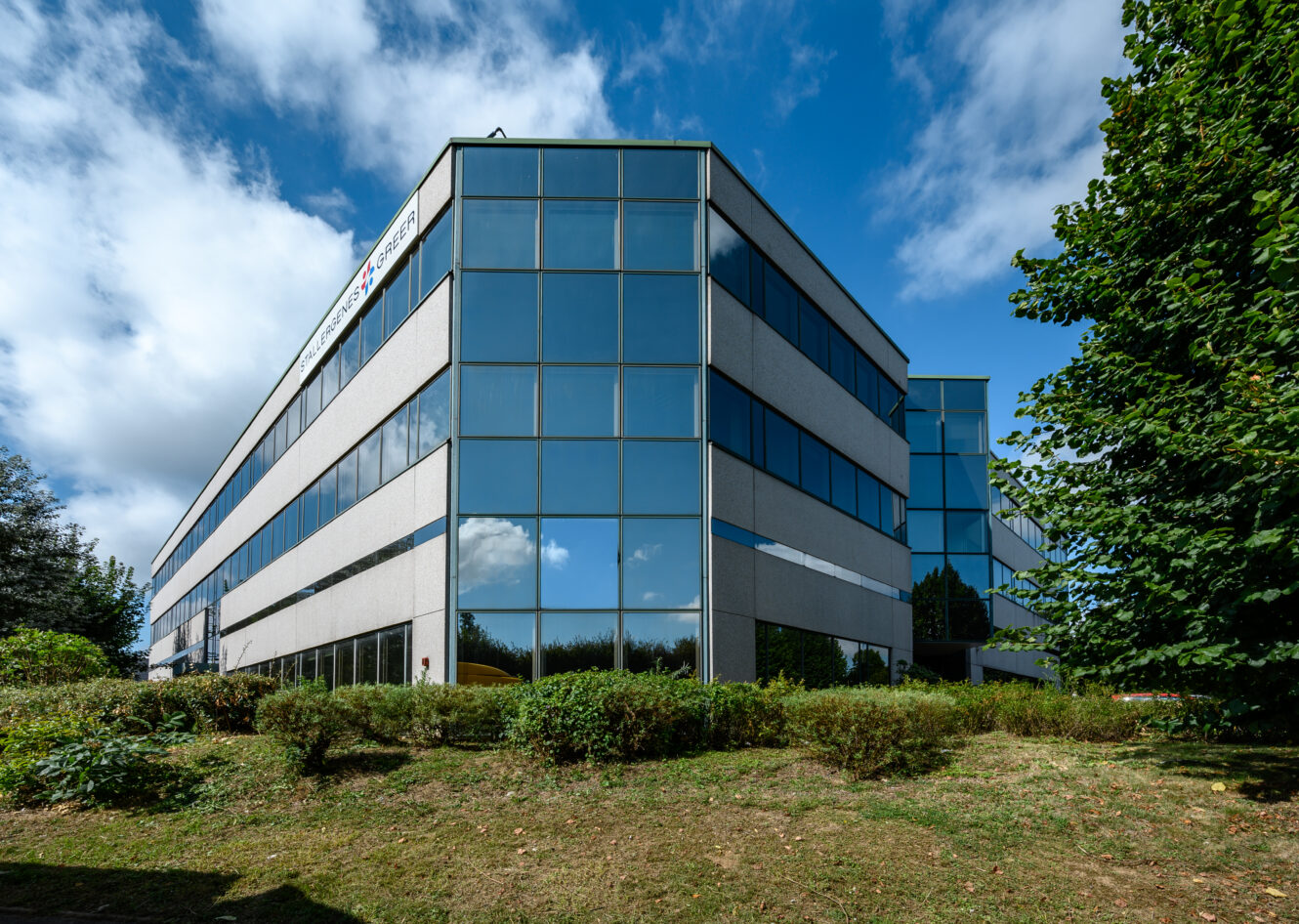 Un immeuble de bureaux moderne de trois étages avec de grandes fenêtres en verre réfléchissant, de la verdure autour et un ciel partiellement nuageux au-dessus.