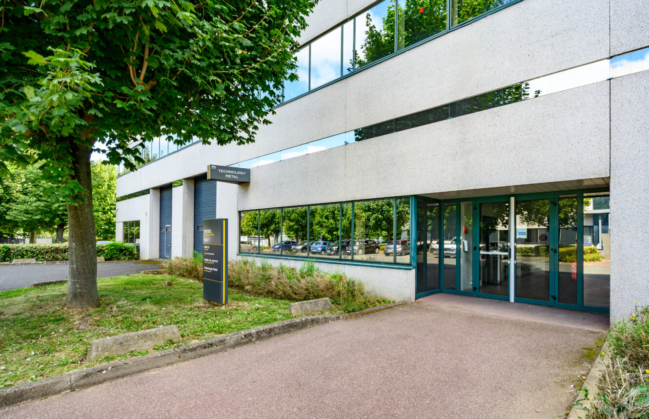 L'entrée d'un immeuble de bureaux moderne avec de grandes fenêtres, un arbre et un panneau au bord du chemin, et des voitures qui se reflètent dans la vitre.