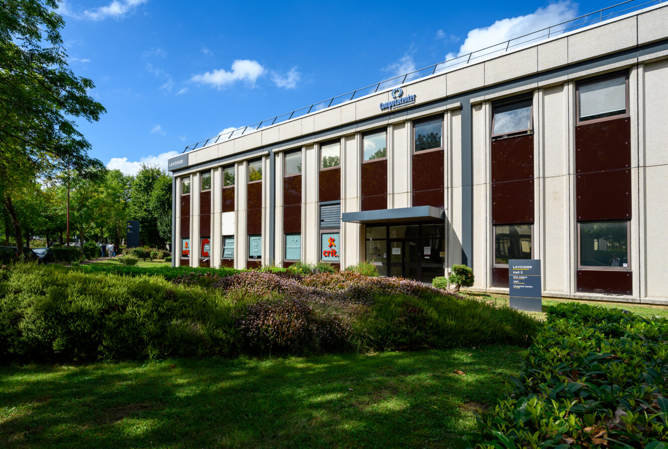 Immeuble de bureaux moderne avec une façade blanche et brune, de grandes fenêtres, un panneau d'entrée, un paysage luxuriant, des arbres et un ciel bleu.