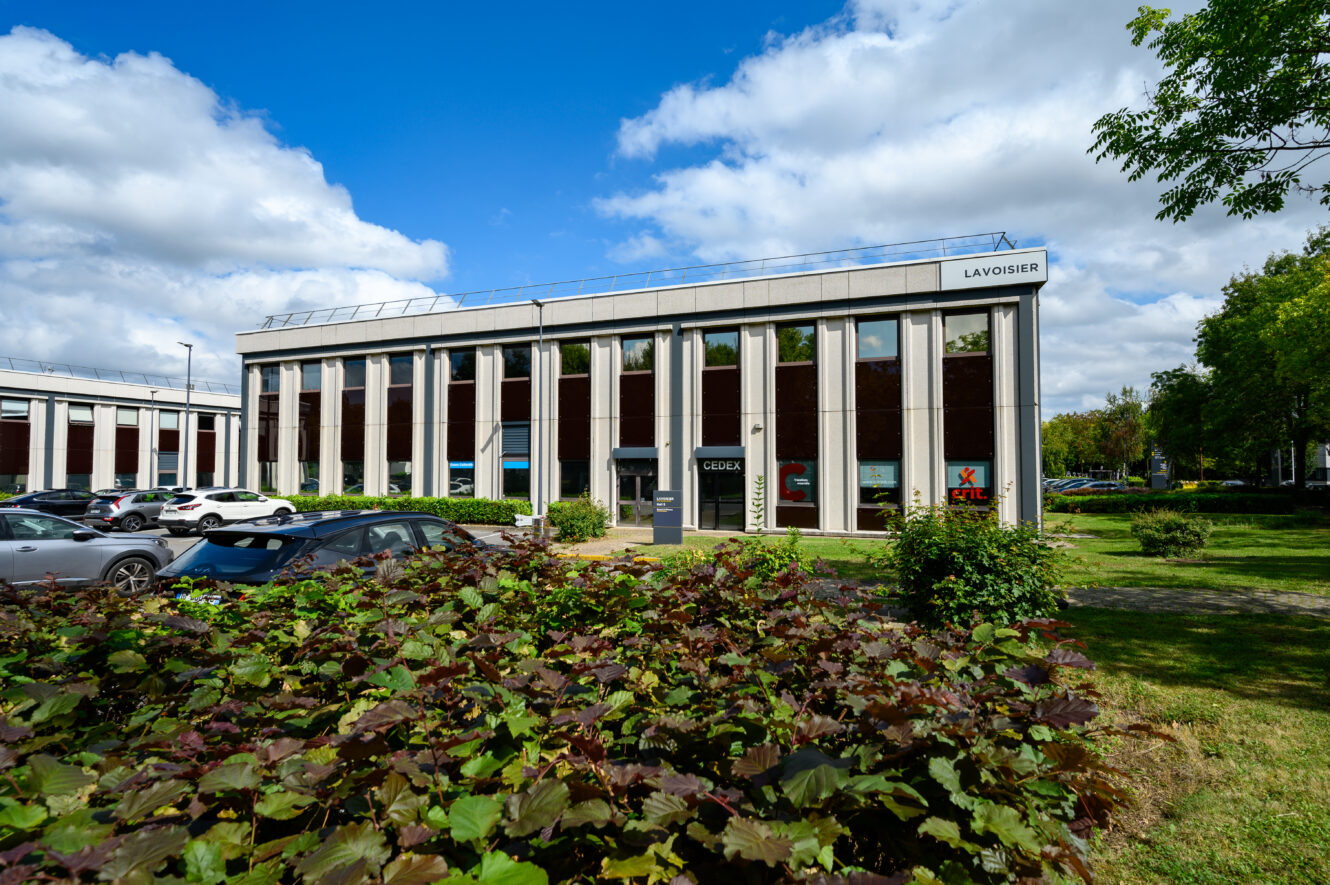 Un immeuble de bureaux moderne de deux étages étiqueté Lavoisier avec de grandes fenêtres, de la verdure et des voitures garées sous un ciel partiellement nuageux.