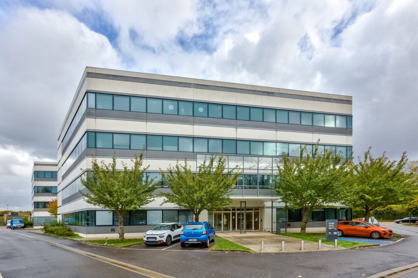 Un immeuble de bureaux moderne de trois étages avec de grandes fenêtres, plusieurs arbres à l'entrée et des voitures garées devant par une journée nuageuse.