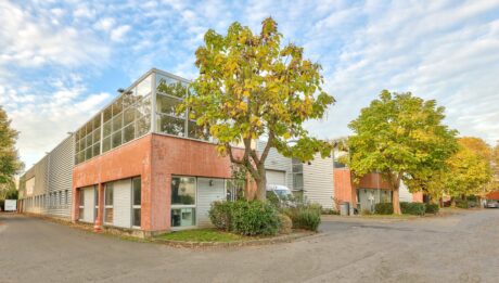 Deux immeubles de bureaux modernes de deux étages avec des extérieurs en brique et en verre sont entourés d'arbres et d'une zone pavée vide sous un ciel partiellement nuageux.