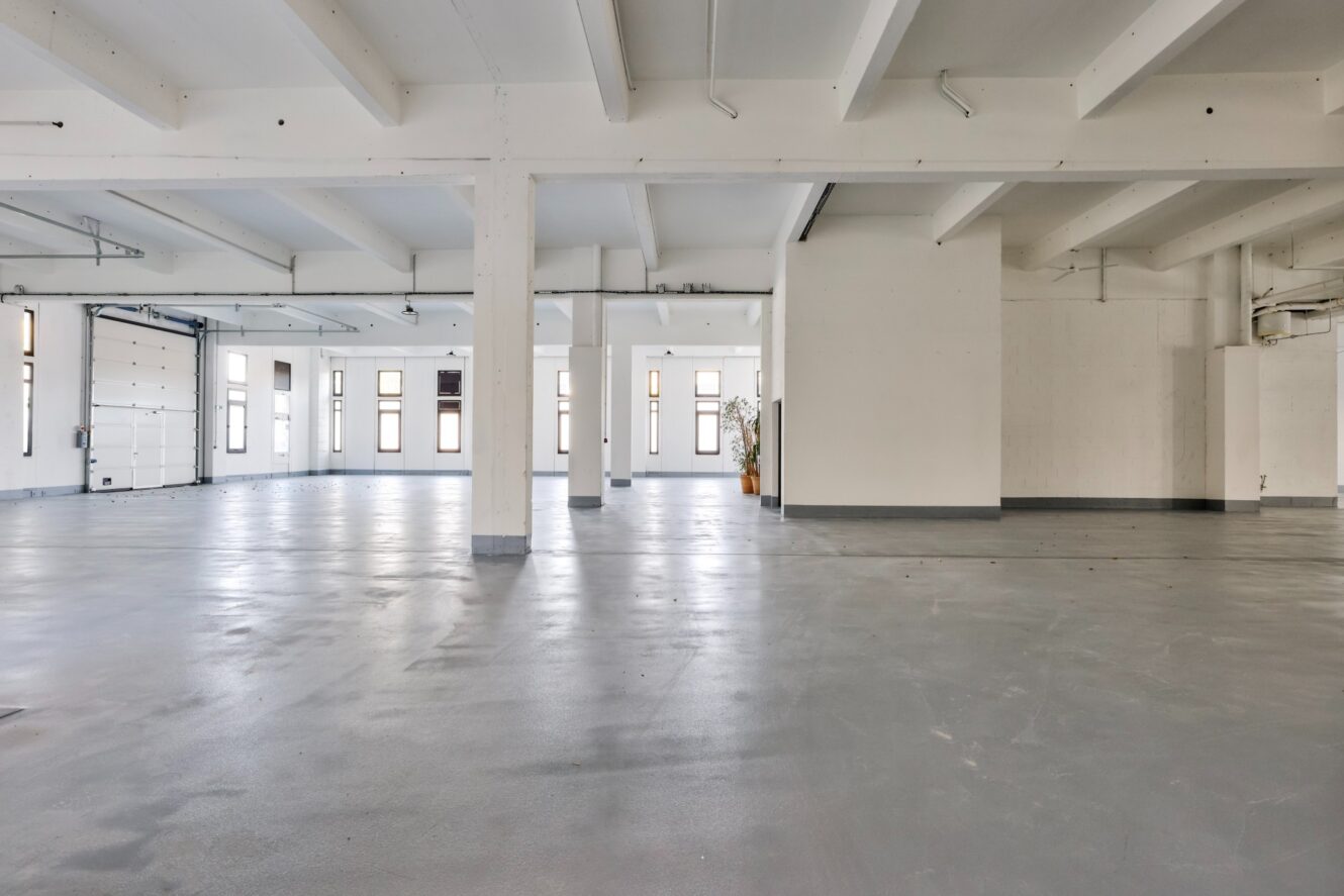 Intérieur industriel spacieux et vide avec des murs blancs, un sol en béton gris, des poutres apparentes et de nombreuses fenêtres laissant entrer la lumière naturelle.