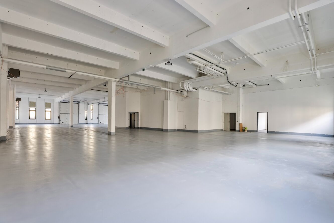 Spacieux entrepôt industriel vide avec des murs blancs, des poutres apparentes et de nombreuses fenêtres et portes. Le sol en béton et les tuyaux du plafond sont visibles dans toute la zone ouverte.