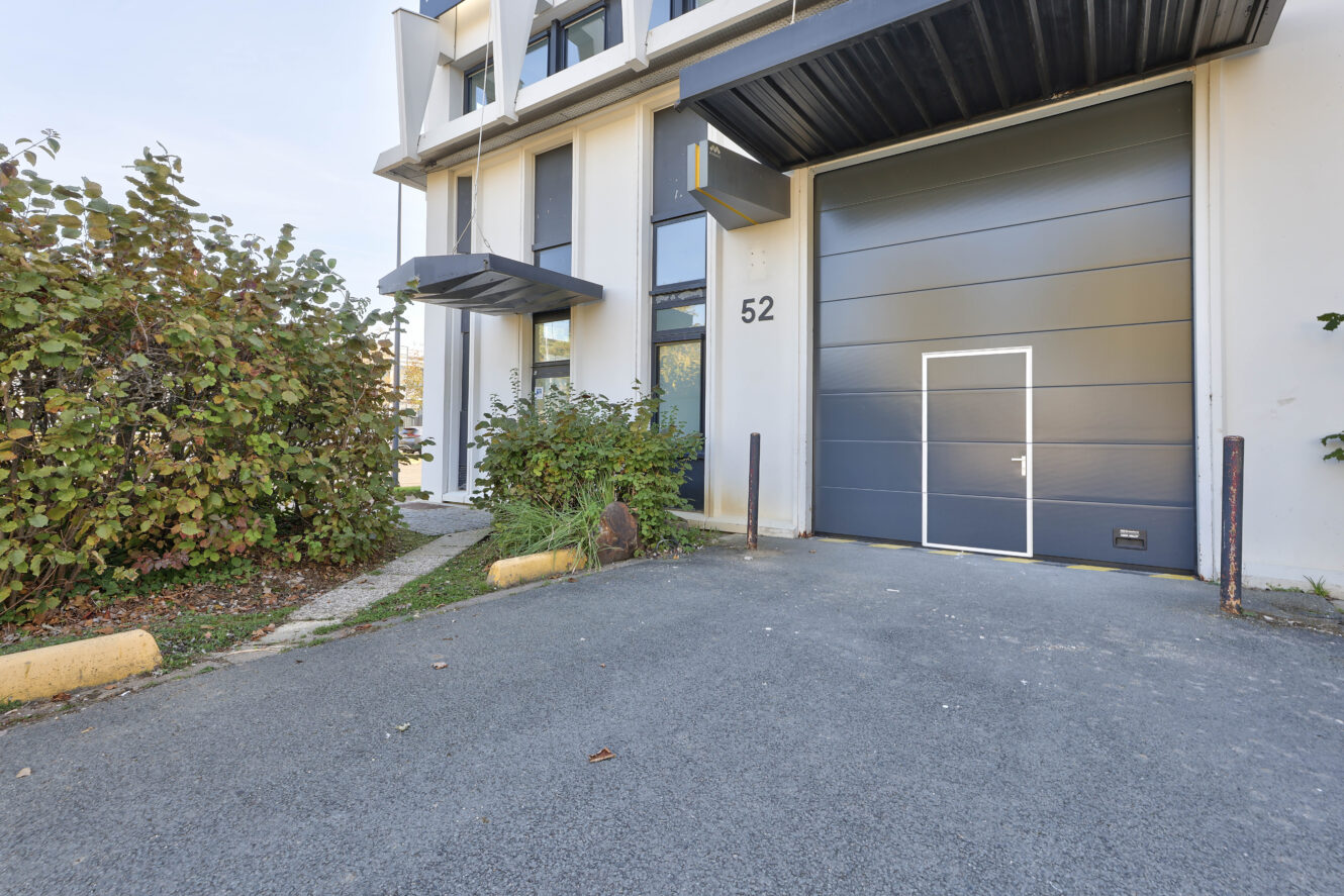 Extérieur d'un bâtiment industriel moderne avec une grande porte de garage grise, une entrée marquée en blanc, le numéro 52, et des buissons à côté de l'allée pavée.