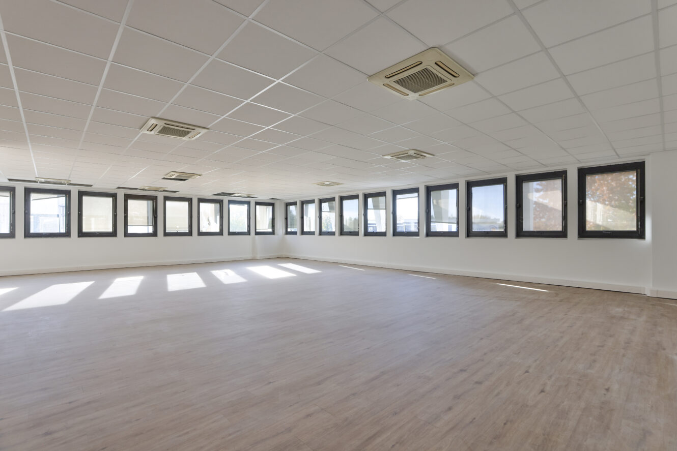 Pièce vide et spacieuse avec un sol en bois clair, des murs blancs, un plafond grillagé avec des bouches d'aération et une rangée de grandes fenêtres rectangulaires laissant entrer la lumière naturelle.