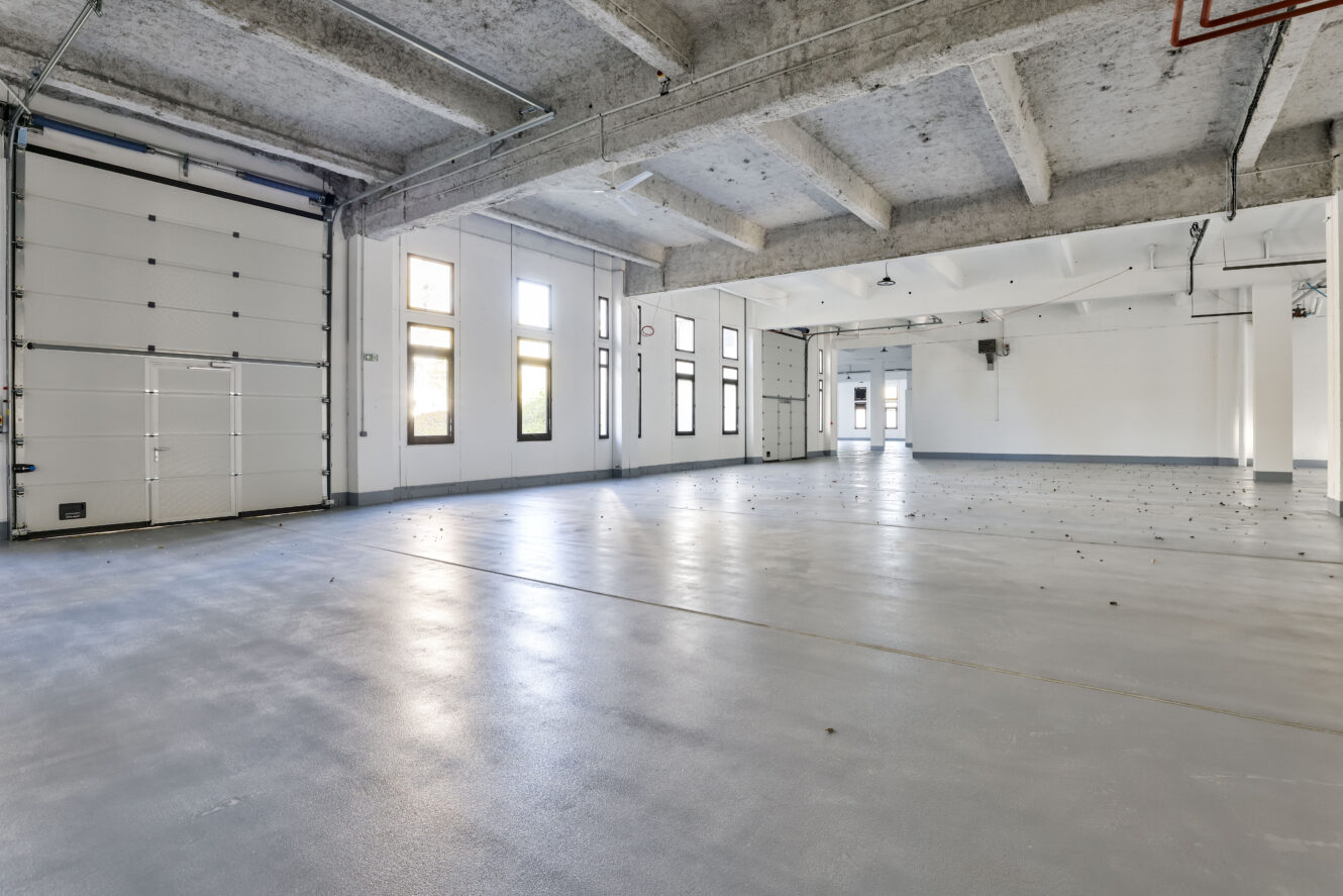 Entrepôt industriel vide avec de hauts plafonds, des poutres apparentes, de grandes fenêtres, une porte de garage enroulable et un sol en béton lisse. La lumière naturelle pénètre par les fenêtres.