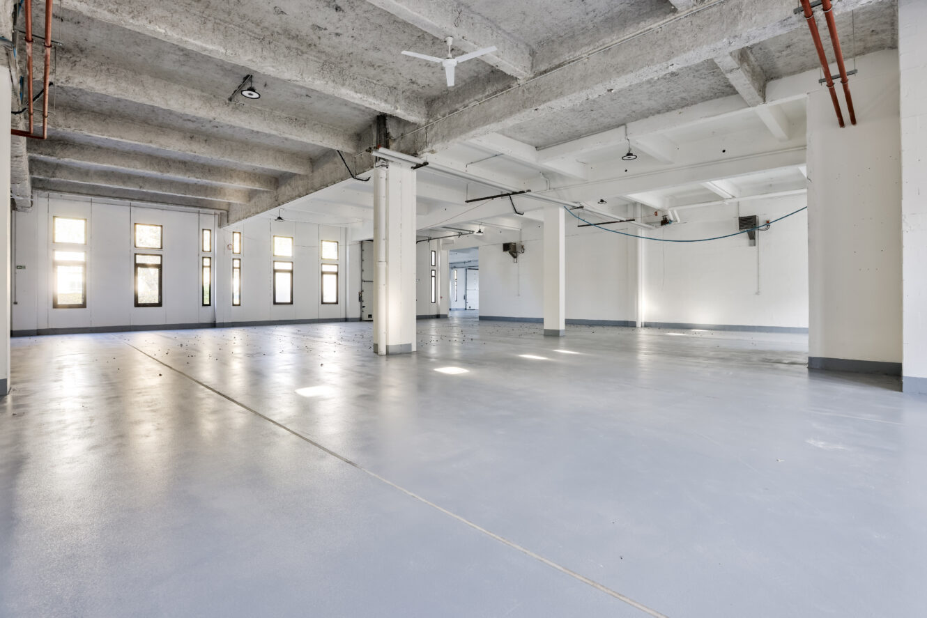 Intérieur industriel spacieux et vide avec des sols en béton, des poutres apparentes, de grandes fenêtres et un éclairage minimal dans un entrepôt ou un espace commercial.