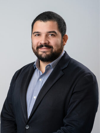 Homme aux cheveux courts et à la barbe foncée, portant une chemise bleu clair à boutons et un blazer foncé, debout sur un fond gris clair uni.