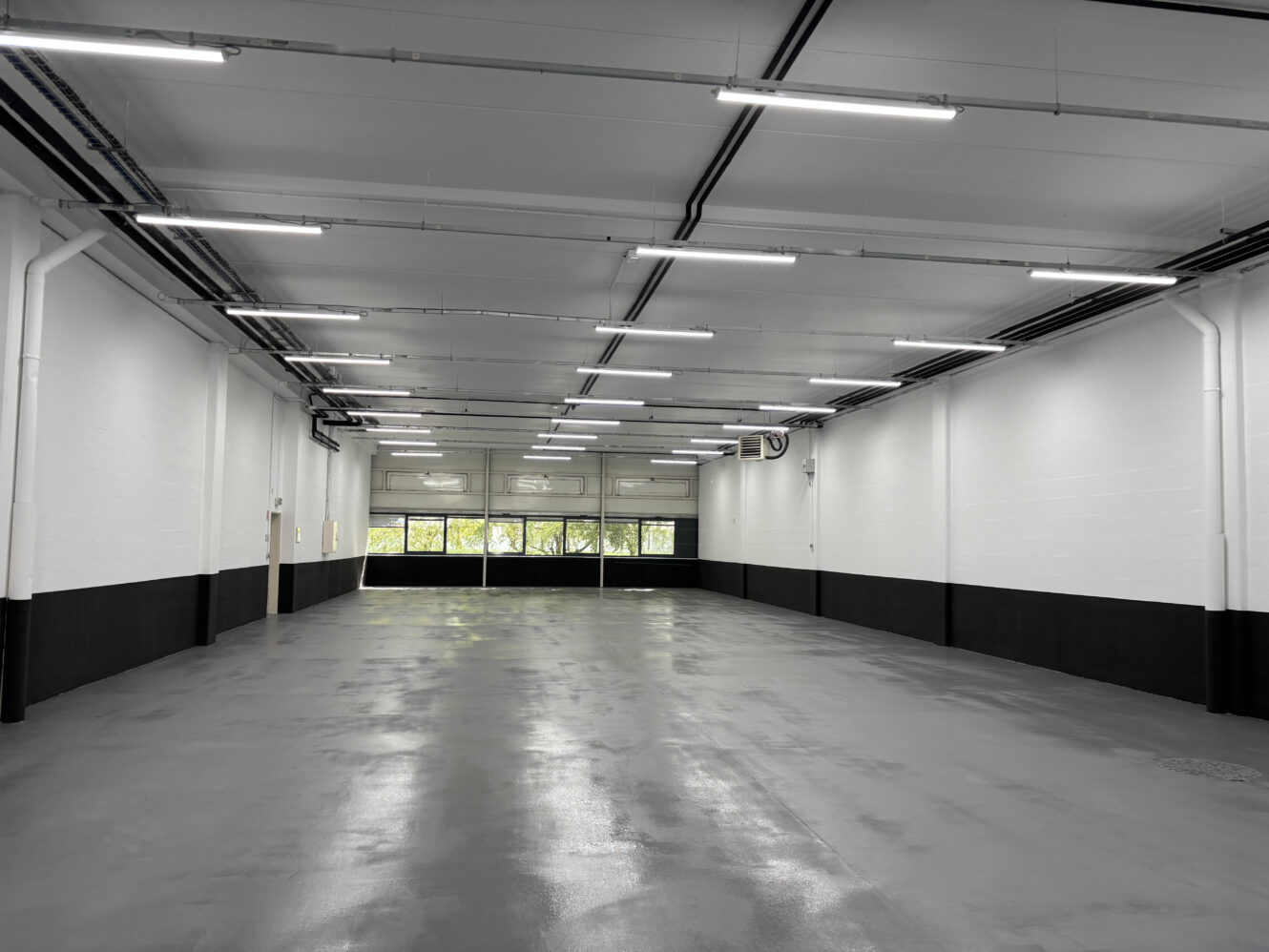 Un garage ou un entrepôt industriel vide avec un sol gris lisse, des murs blancs, des garnitures noires et un éclairage fluorescent au plafond. De grandes fenêtres sont visibles au fond.