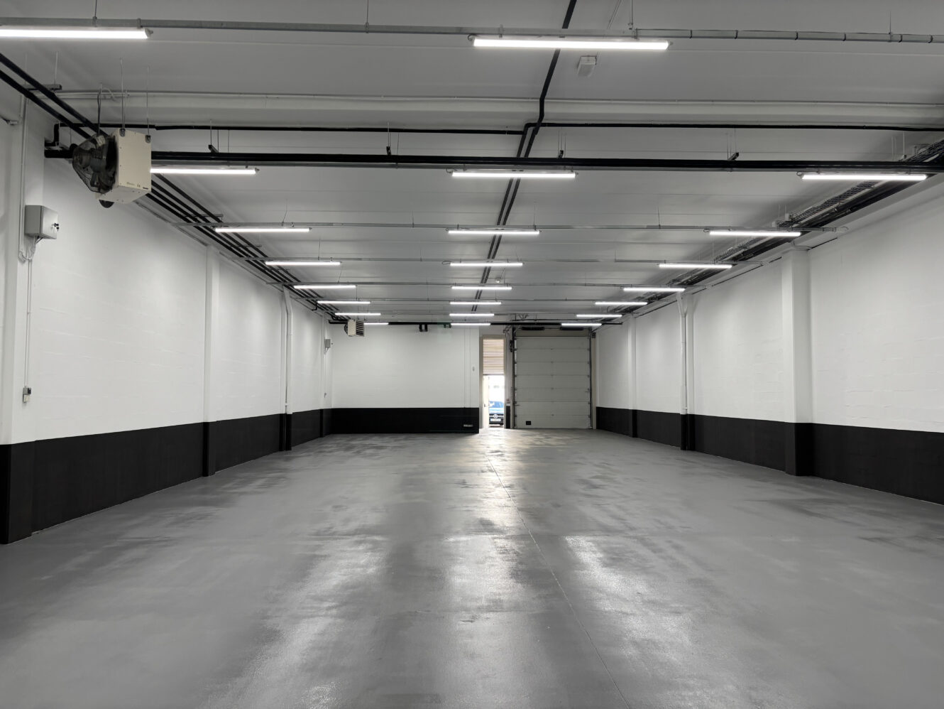 Entrepôt ou garage industriel vide et bien éclairé, avec un sol en béton poli, des murs blancs, des garnitures noires, des plafonniers, des tuyaux apparents et une porte de garage partiellement ouverte à l'extrémité.