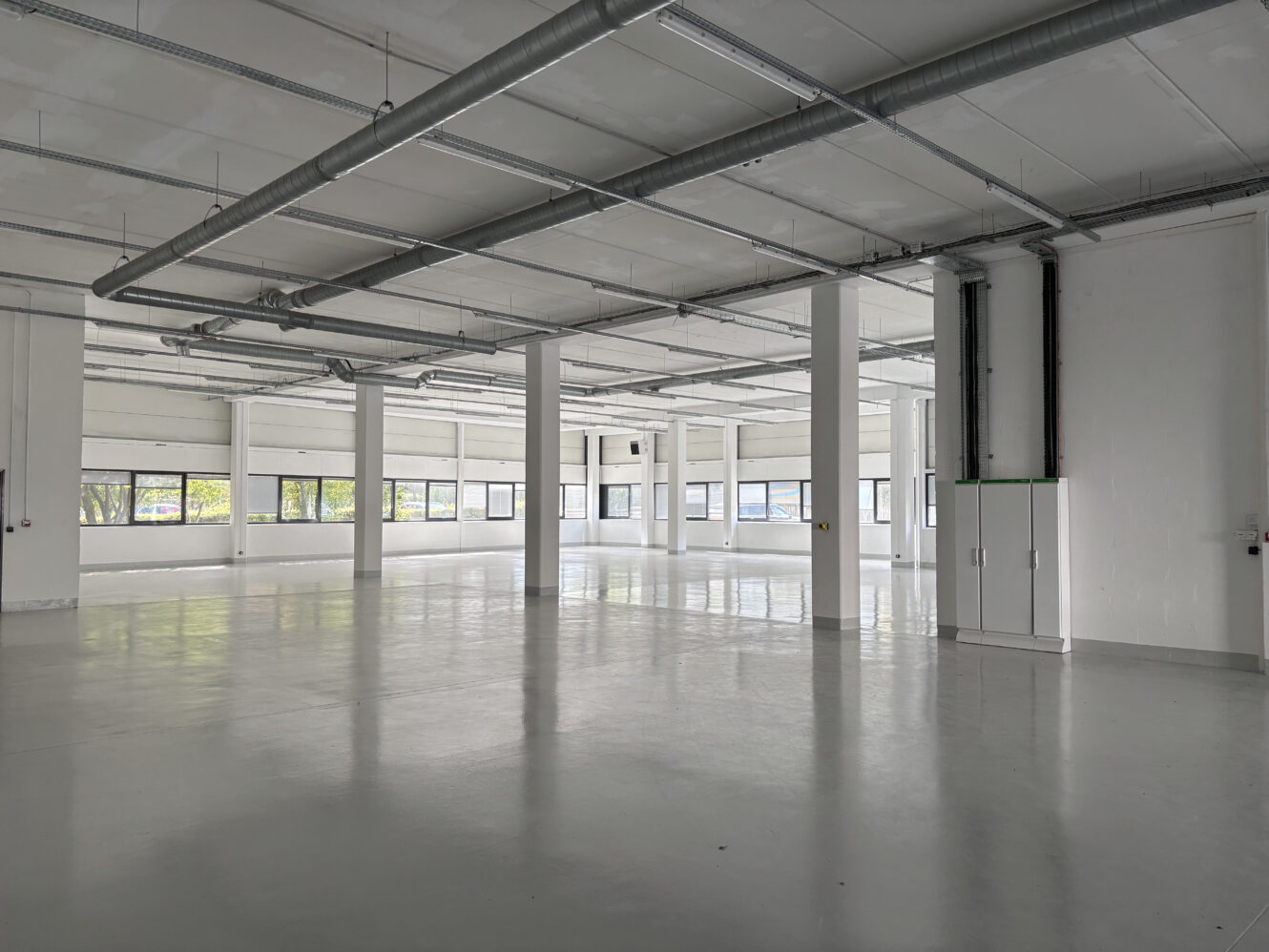 Pièce industrielle spacieuse et vide avec des murs blancs, de grandes fenêtres, des conduits apparents et un sol en béton poli.