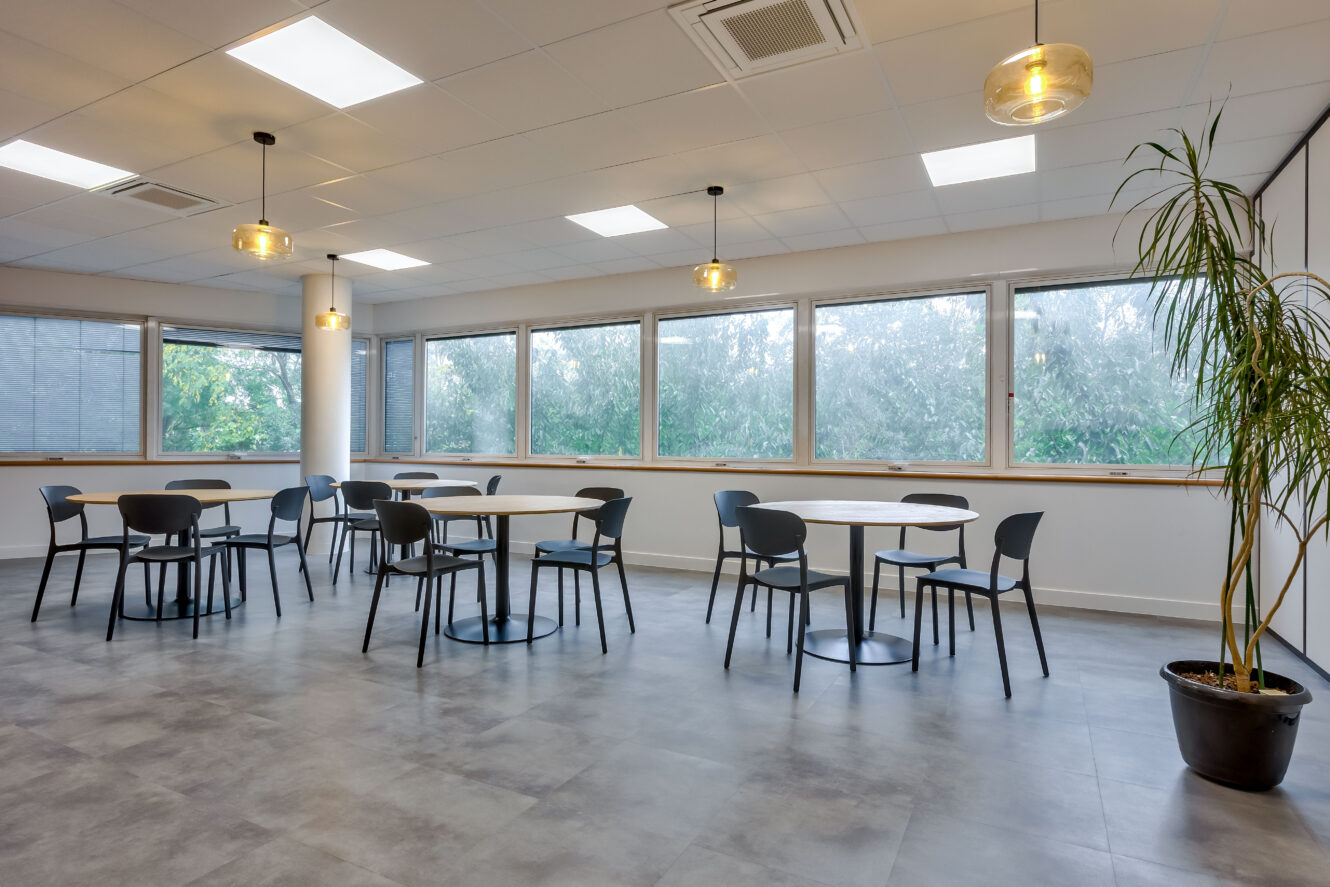 Une cafétéria moderne et vide avec des tables rondes, des chaises noires, de grandes fenêtres, des plafonniers et une plante en pot dans un coin.