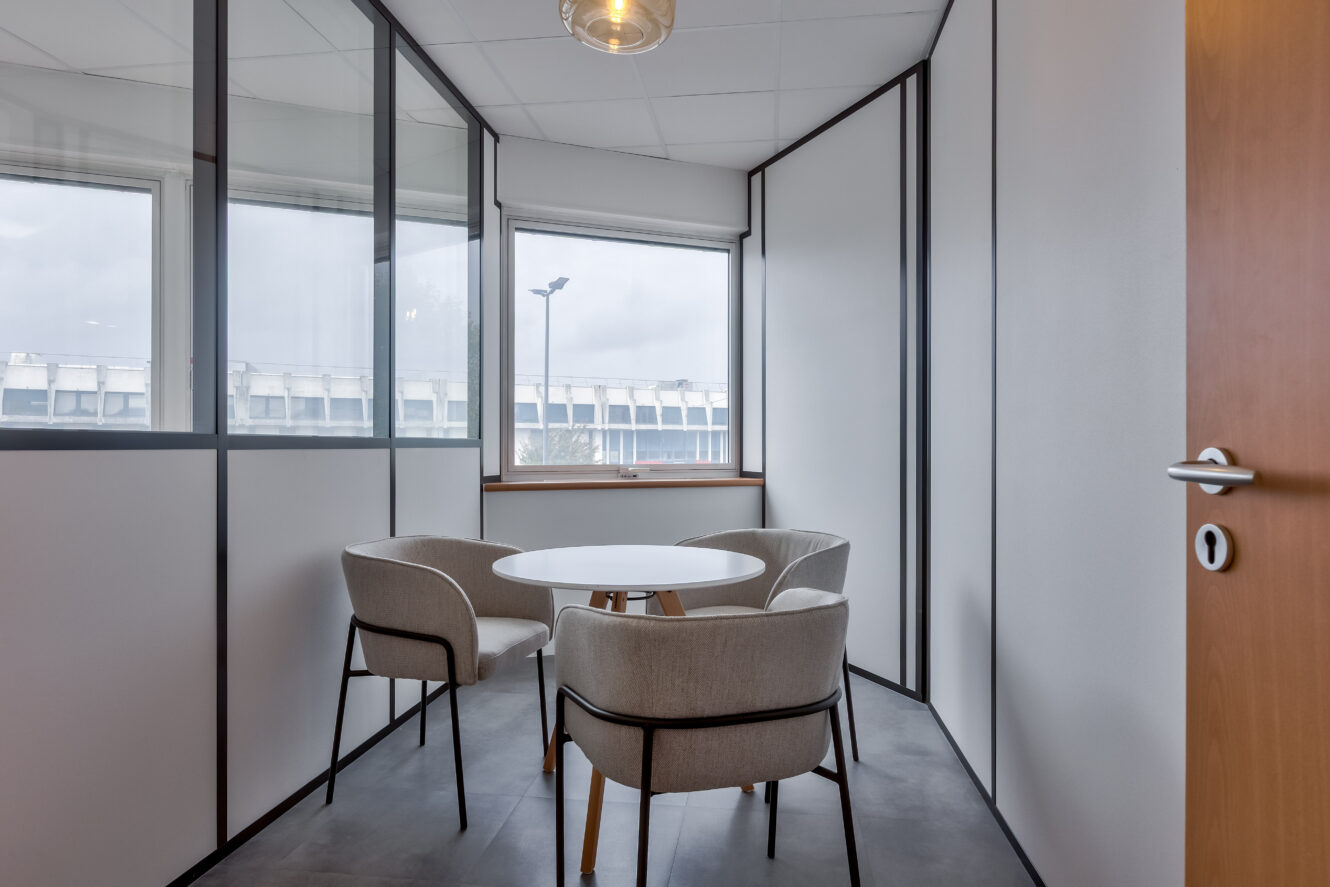 Petite salle de conférence avec une table ronde, quatre chaises beiges, des cloisons en verre et de grandes fenêtres laissant entrer la lumière naturelle.