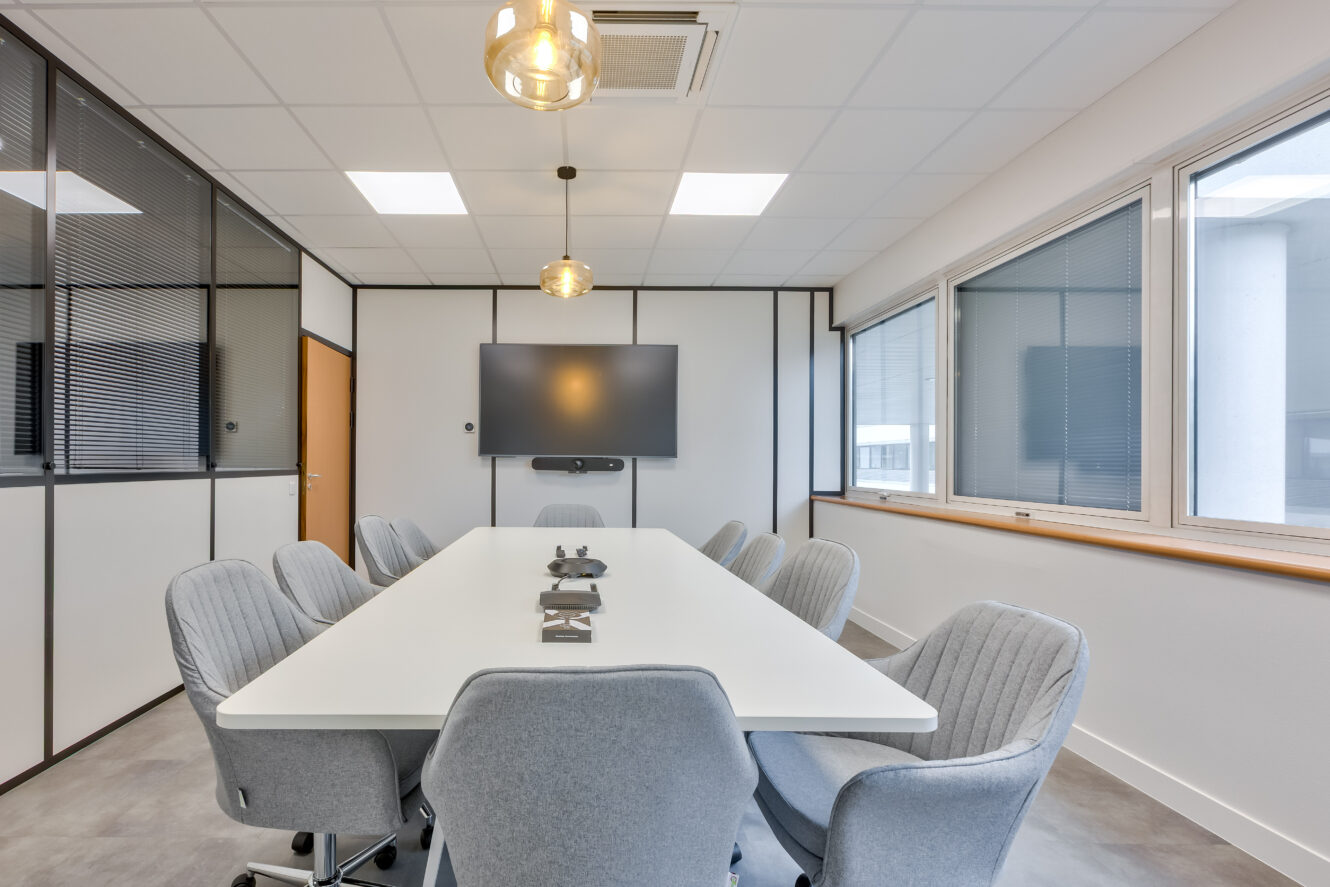Une salle de conférence moderne avec une longue table blanche, huit chaises grises, un grand écran mural, des plafonniers et des fenêtres avec des stores.