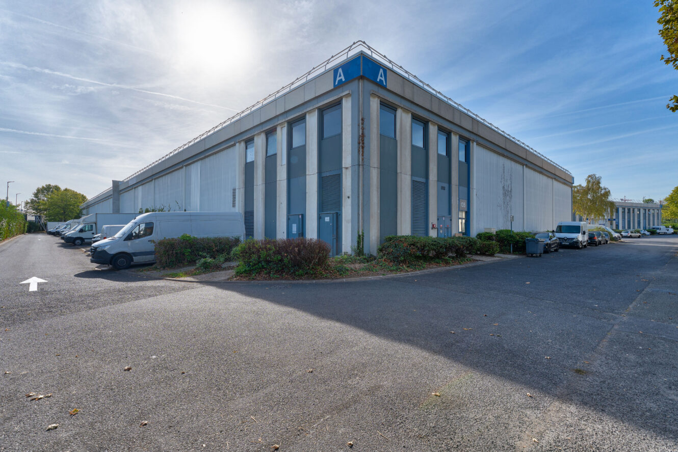 Un grand entrepôt industriel devant lequel sont garées plusieurs camionnettes blanches, par une journée ensoleillée. Le bâtiment est orné de panneaux bleus en forme de « A » à ses angles, et un parking pavé s'étend au premier plan.