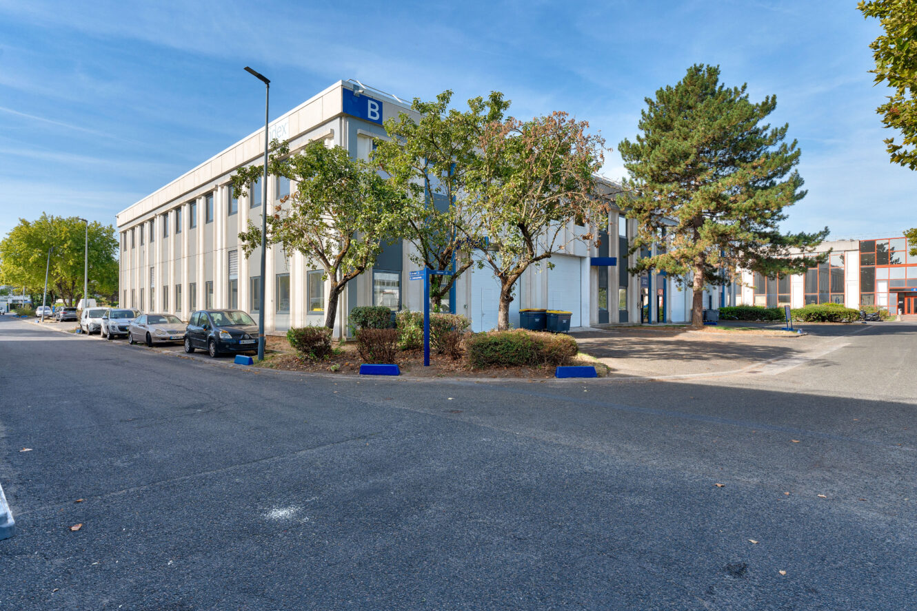 Un immeuble de bureaux de deux étages, orné d'une enseigne bleue en forme de « B », entouré d'arbres et de voitures garées le long de la rue, par une journée ensoleillée.