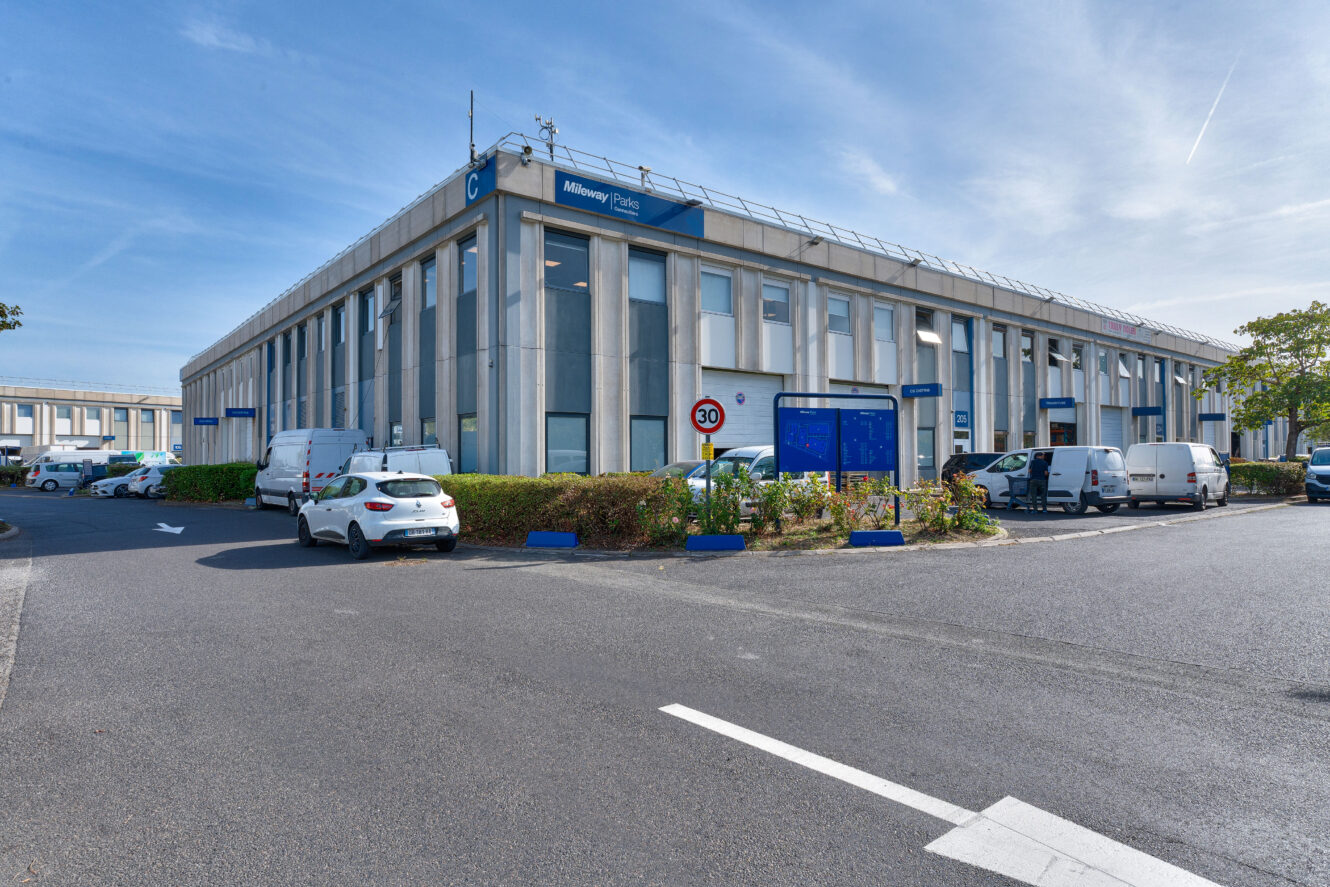 Bâtiment industriel à deux étages avec plusieurs voitures et camionnettes garées, une enseigne bleue près de l'entrée et un panneau de limitation de vitesse au premier plan, sous un ciel dégagé.
