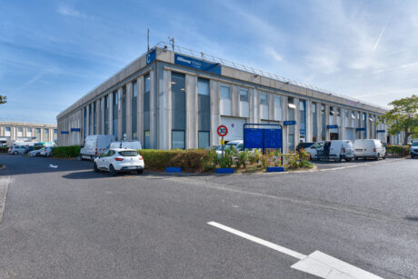 Bâtiment industriel à deux étages avec plusieurs voitures et camionnettes garées, une enseigne bleue près de l'entrée et un panneau de limitation de vitesse au premier plan, sous un ciel dégagé.
