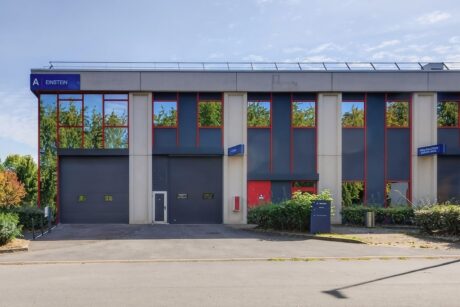 Un immeuble commercial moderne de deux étages, doté de grandes baies vitrées, d'une façade grise et d'enseignes bleues portant l'inscription « Einstein » ; entouré de verdure et d'arbres dont les reflets se dessinent sur les vitres.