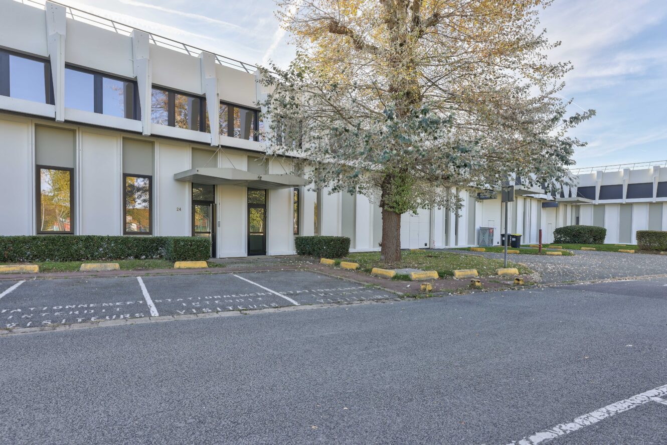 Un immeuble de bureaux moderne de deux étages avec de grandes fenêtres et un parking devant, partiellement ombragé par un arbre. Plusieurs places de parking vides sont visibles.