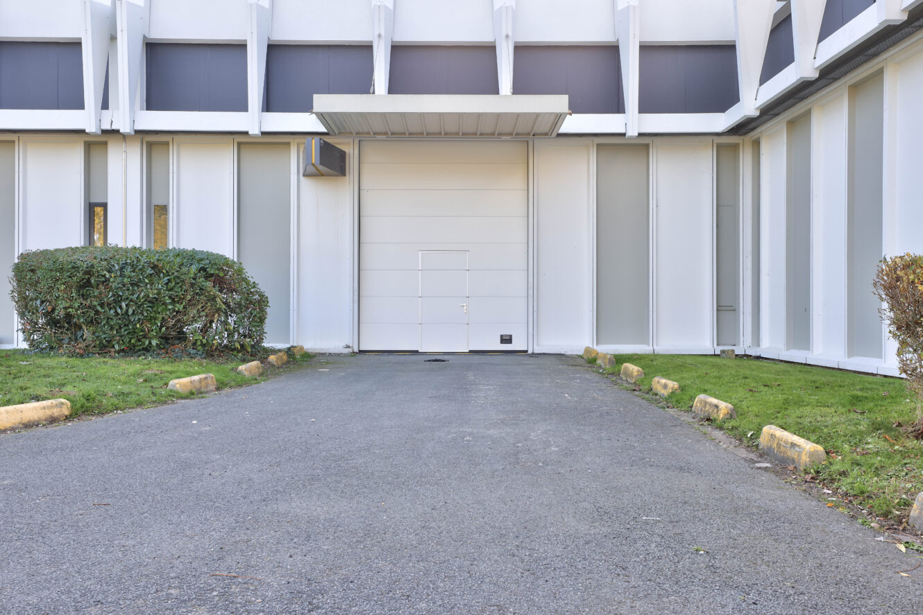 Une allée mène à une grande porte blanche située dans un bâtiment moderne blanc, flanqué de buissons taillés et de barrières de parking de chaque côté.