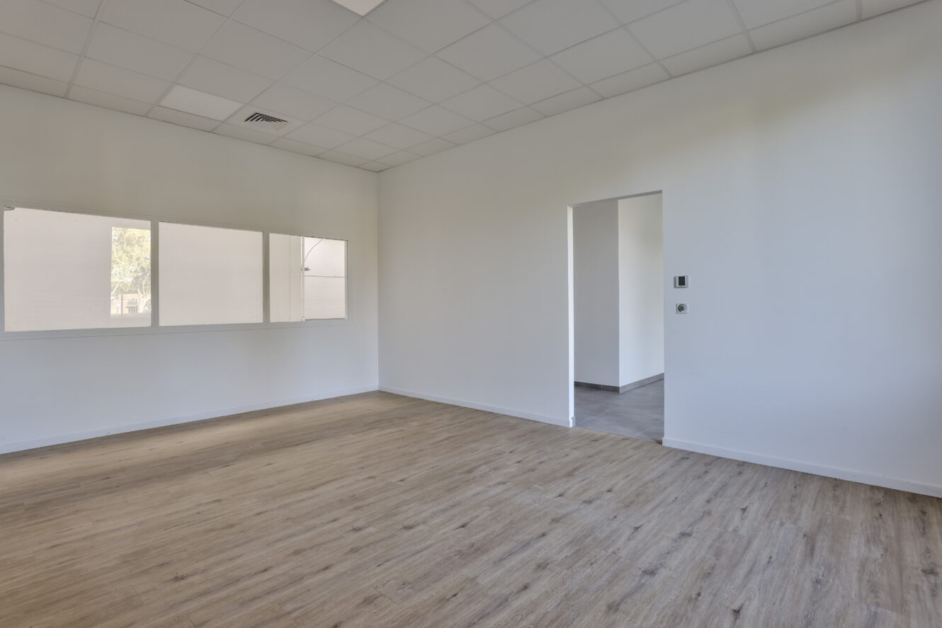 Pièce vide avec un sol en bois clair, des murs blancs, une porte menant à une autre zone et trois fenêtres rectangulaires sur un mur.