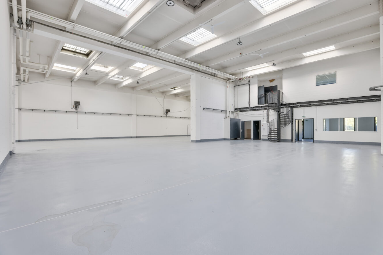 Spacieux entrepôt industriel vide avec des murs blancs, de hauts plafonds, des puits de lumière, un sol gris et un escalier métallique menant à un bureau supérieur.