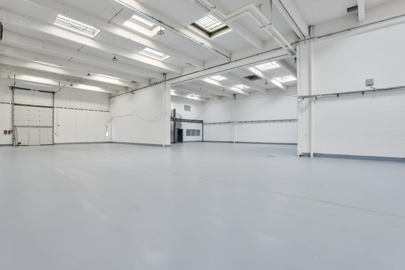 Spacieux entrepôt industriel vide avec des murs blancs, de hauts plafonds, des puits de lumière et un sol en béton. Un escalier métallique et une porte roulante fermée sont visibles à l'arrière-plan.