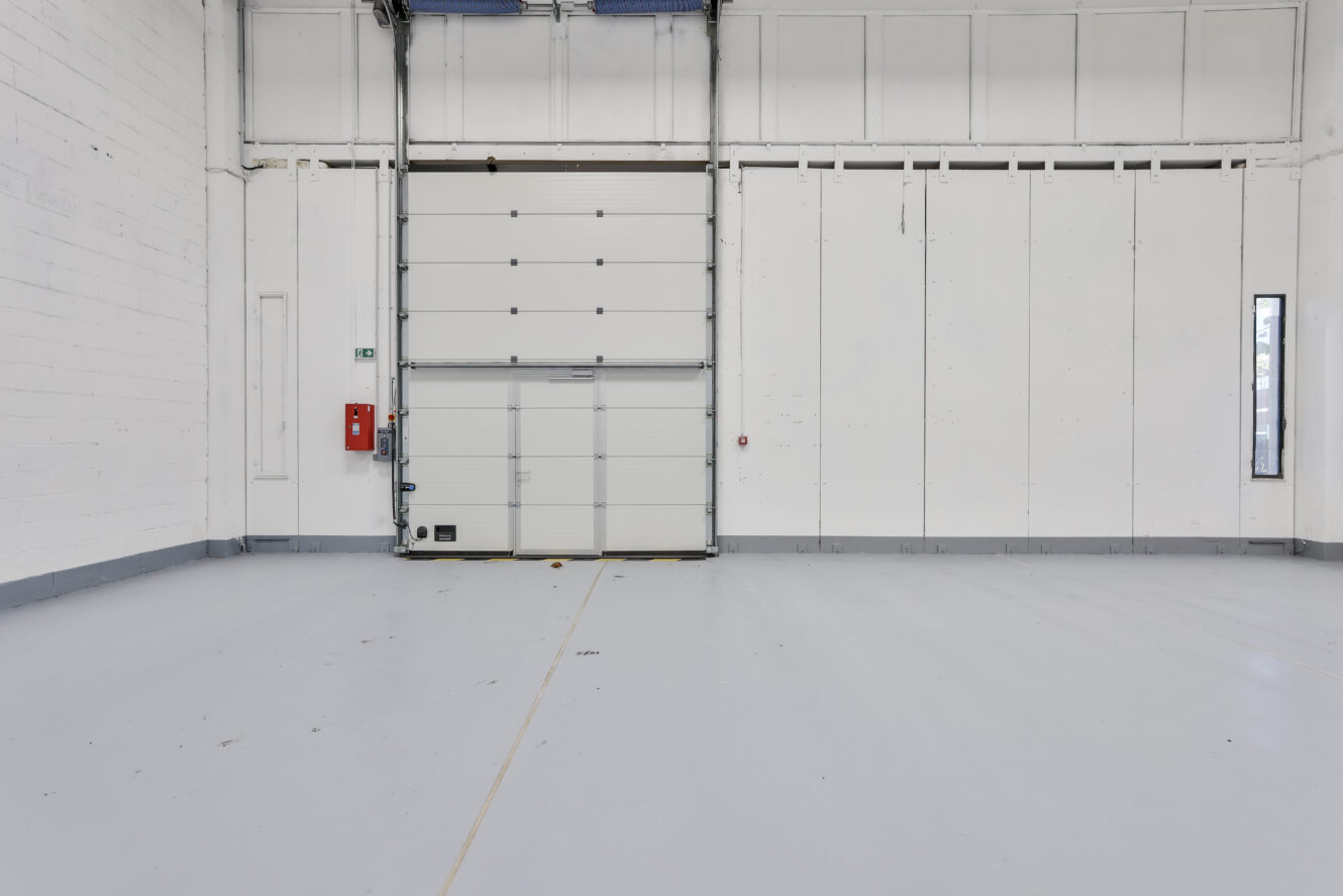 Entrepôt industriel vide avec une porte roulante blanche, une alarme incendie et un panneau de sortie de secours sur le mur gauche ; sol lisse gris clair et murs blancs.
