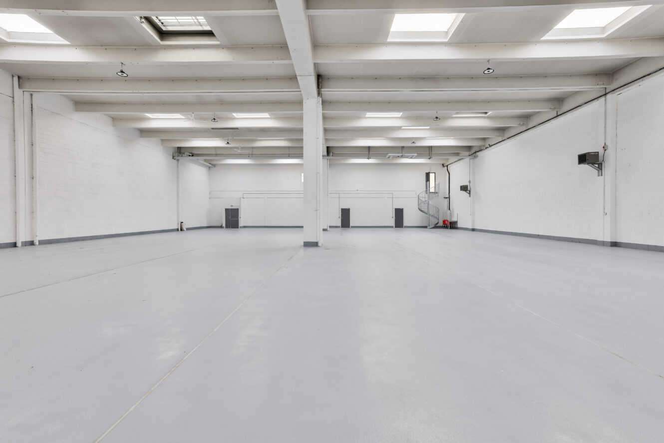 Spacieux entrepôt vide avec des murs blancs, un haut plafond, des puits de lumière et des portes grises. Le sol est lisse et propre, et la zone est bien éclairée par la lumière naturelle.