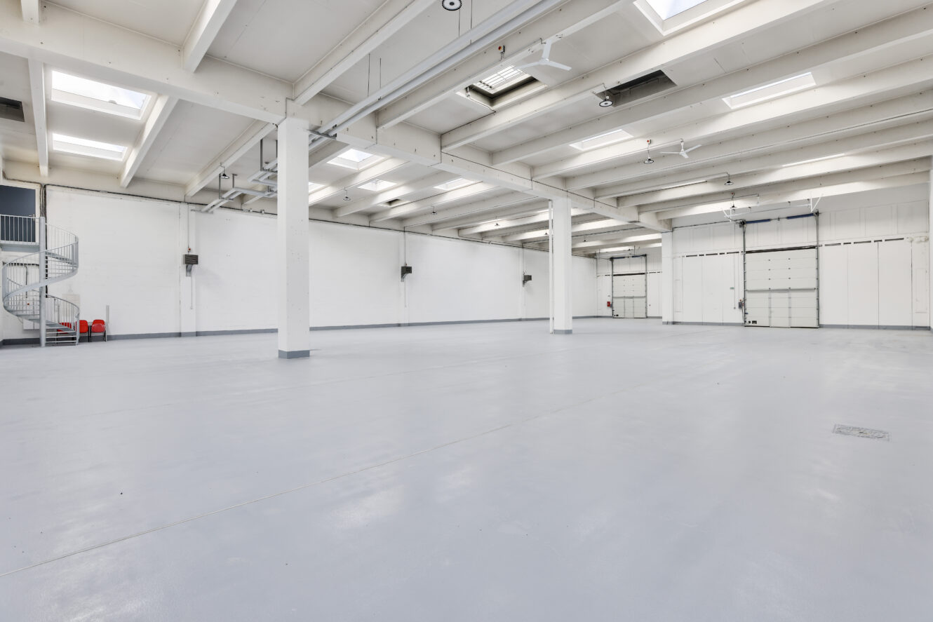 Grand entrepôt industriel vide avec des murs blancs, des lucarnes, des poutres apparentes et un escalier en colimaçon dans le coin.