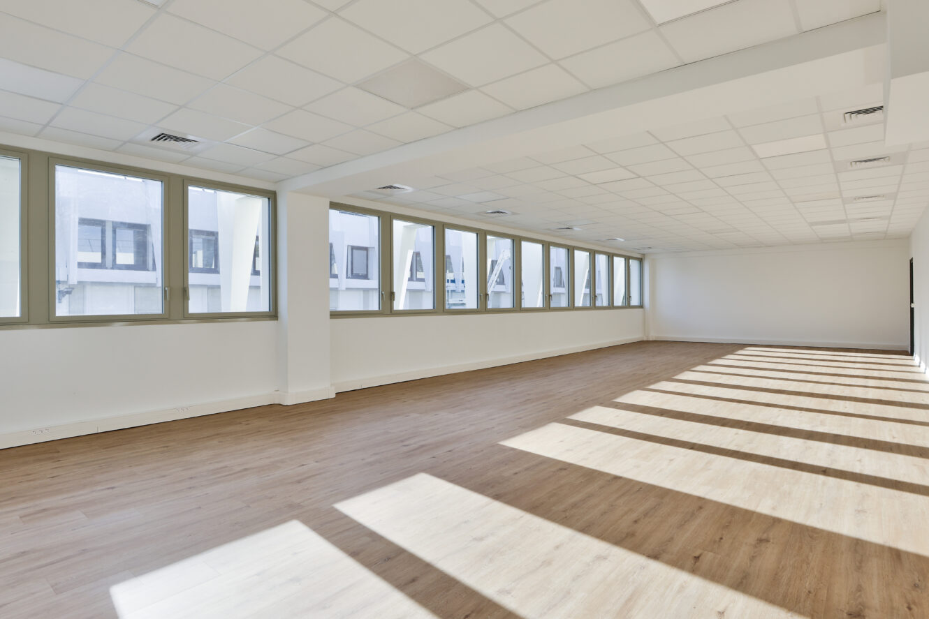 Pièce vide et spacieuse avec de grandes fenêtres, des murs blancs, un plancher en bois et un plafond quadrillé. La lumière du soleil projette de longues ombres sur le sol depuis le côté droit.