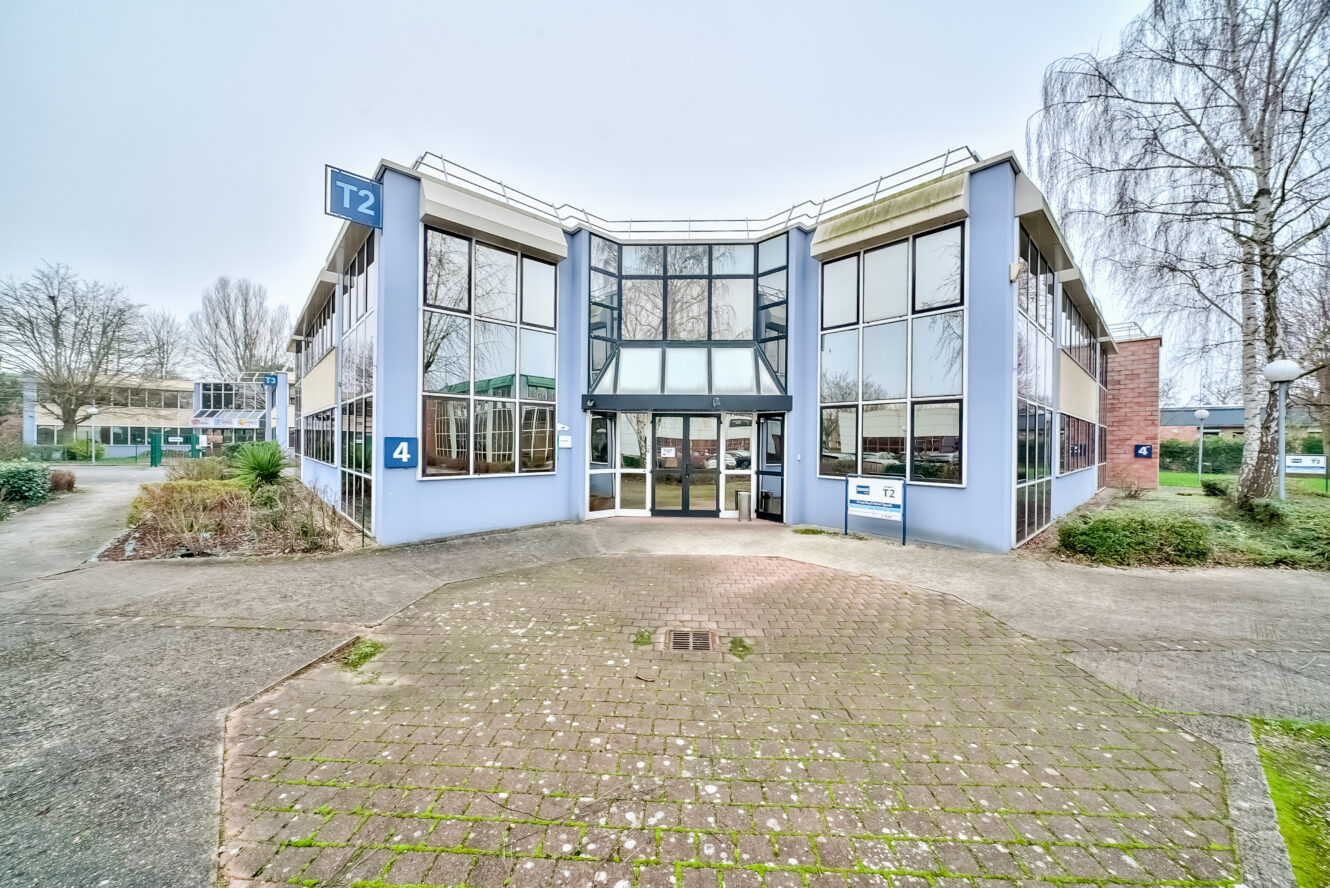 Immeuble de bureaux moderne de deux étages avec de grandes fenêtres, étiqueté T2, et entouré d'une allée pavée, de gazon et de quelques arbres.
