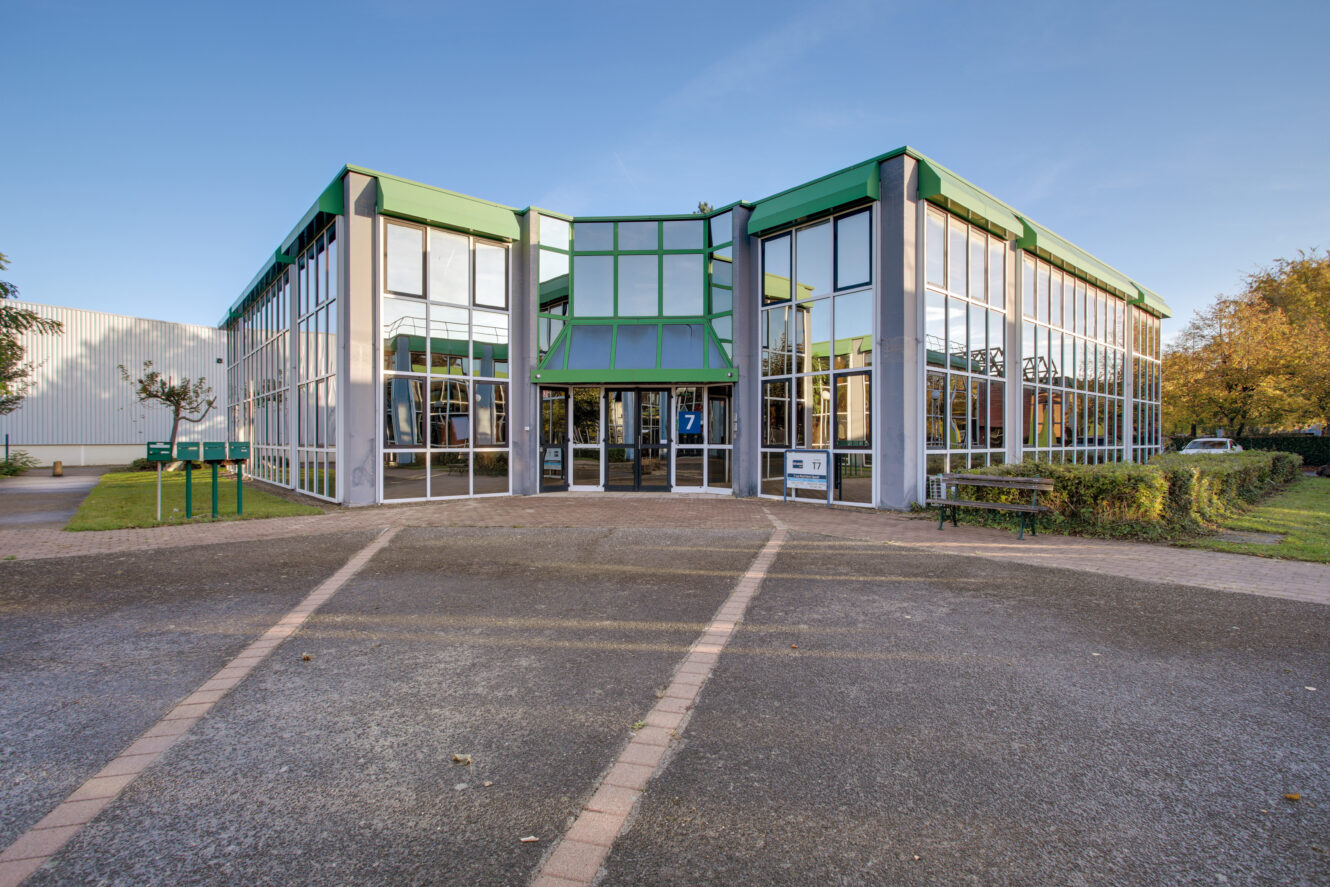 Immeuble de bureaux moderne de deux étages avec de grandes baies vitrées et des bordures vertes, entouré d'une allée pavée et d'un peu de verdure.