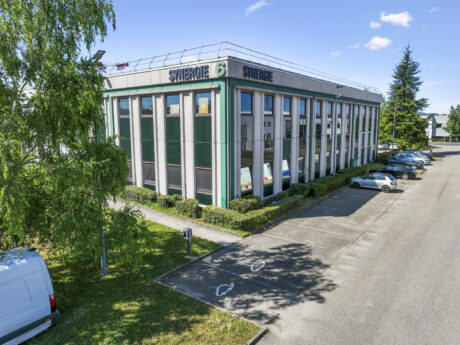 Un immeuble de bureaux moderne portant le nom de "SYNERGIE" avec de grandes fenêtres, un parking adjacent, plusieurs voitures garées et la verdure environnante sous un ciel bleu.