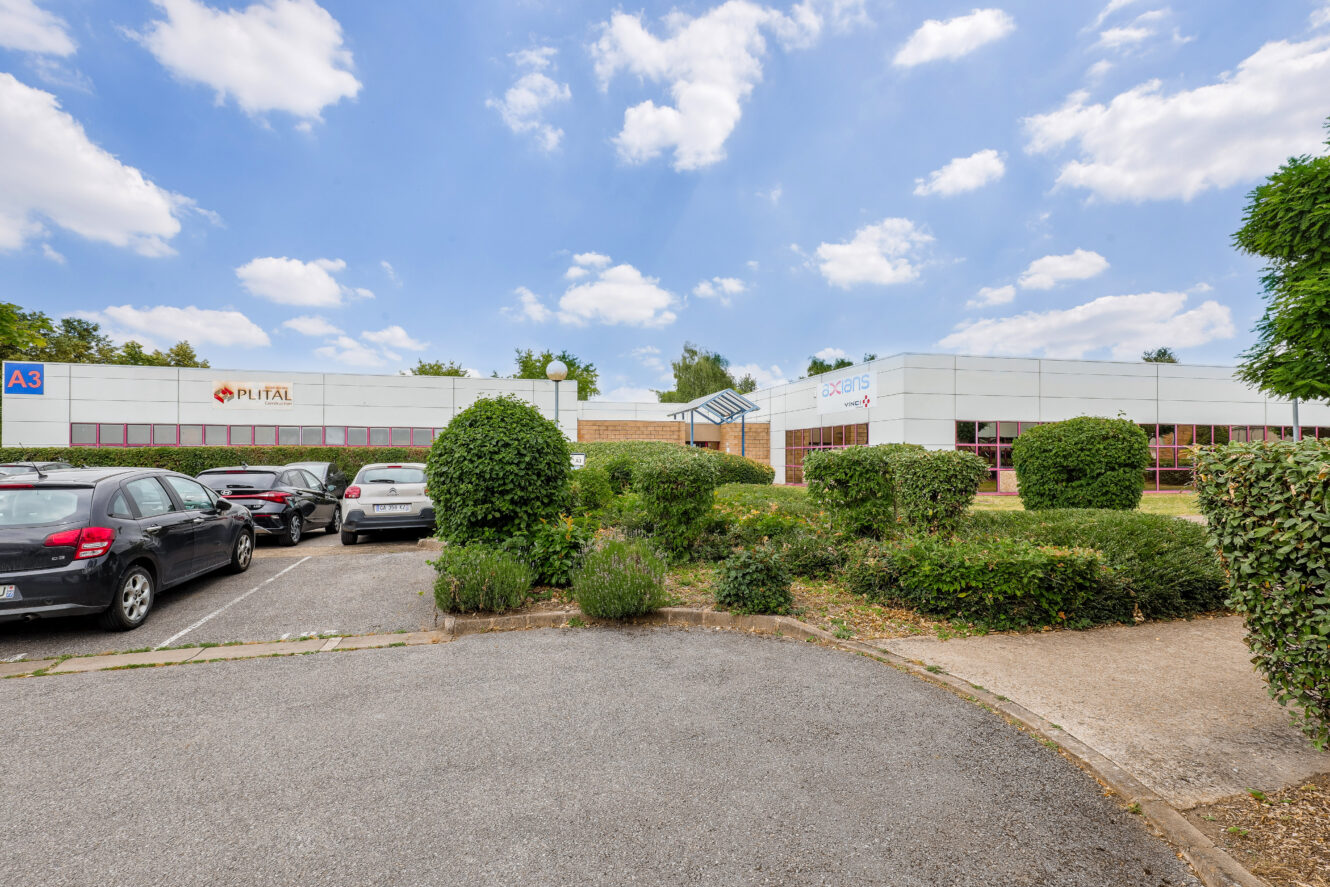 Un parking avec plusieurs voitures devant un bâtiment commercial bas étiqueté Pufital et Jacko S par une journée partiellement nuageuse. Des arbustes et de la verdure entourent la zone.