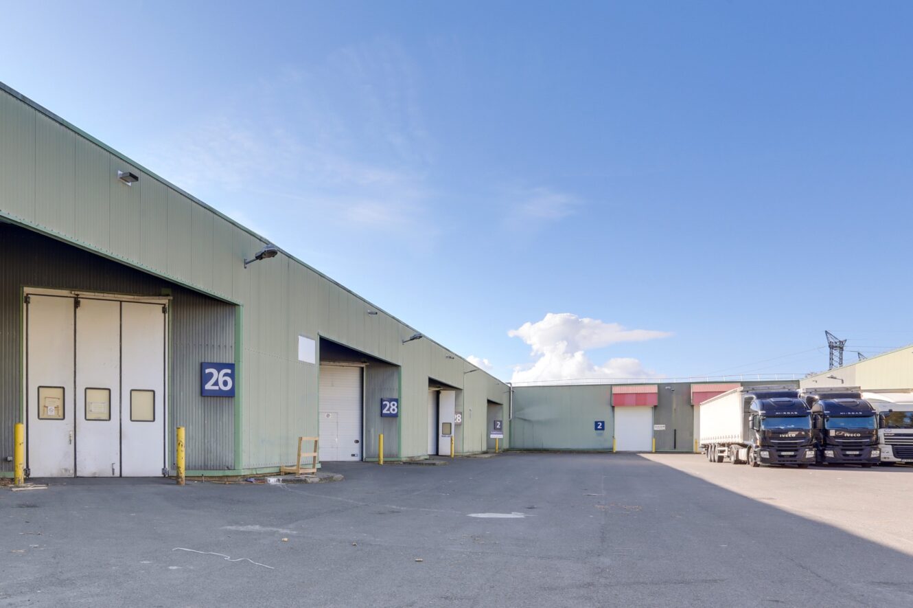Extérieur d'un entrepôt industriel avec des quais de chargement numérotés, dont les quais 26 et 28, et plusieurs camions stationnés par temps clair.