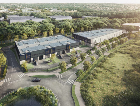 Vue aérienne de bâtiments industriels modernes entourés d'arbres, de parkings et de routes, avec une zone herbeuse et un étang à proximité.