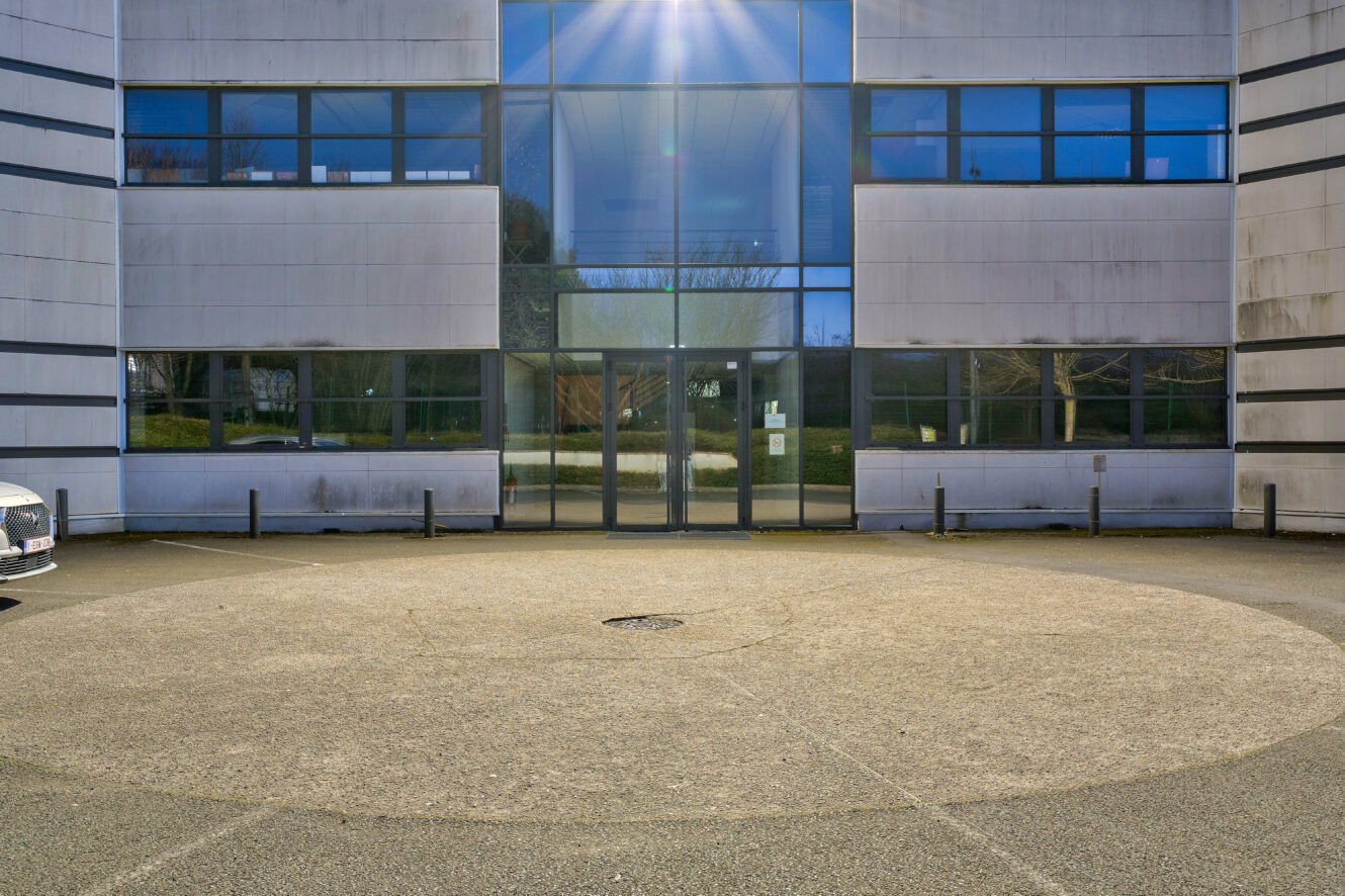 L'image montre l'extérieur d'un bâtiment moderne avec de grandes fenêtres en verre, des portes à double vitrage et une allée circulaire pavée à l'avant.