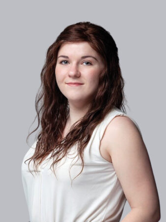 Une femme aux longs cheveux bruns ondulés, vêtue d'un haut blanc sans manches, se tient sur un fond gris clair uni.