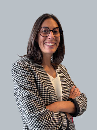 Une femme aux cheveux bruns raides et aux lunettes sourit, les bras croisés, vêtue d'un blazer pied-de-poule noir et blanc sur un haut blanc, sur un fond gris uni.