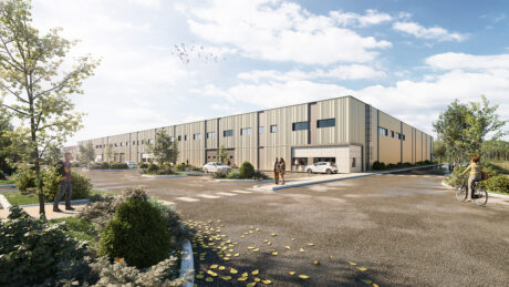 Un entrepôt industriel moderne avec un revêtement beige, de grandes fenêtres et plusieurs voitures garées à l'extérieur ; des gens marchent et font du vélo à proximité sous un ciel partiellement nuageux.