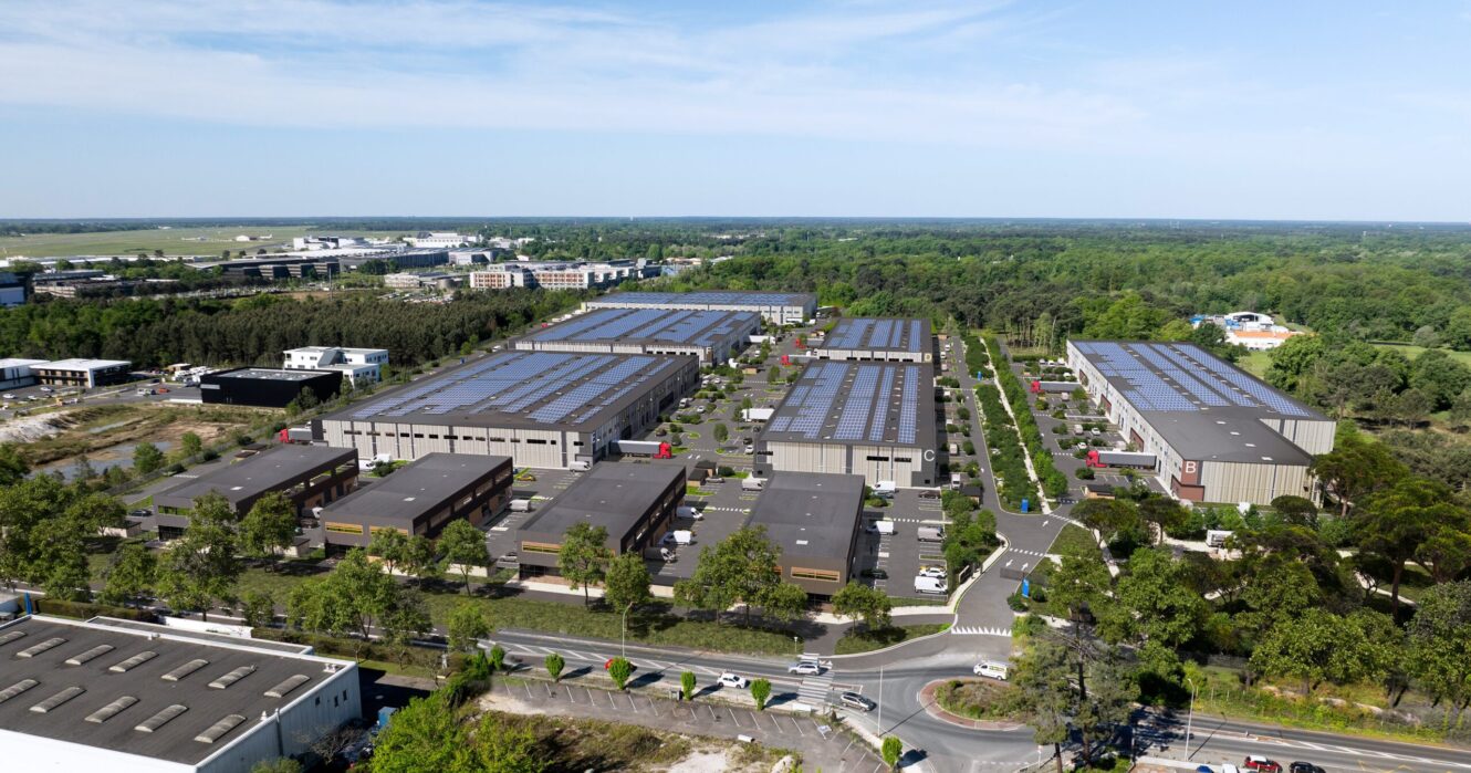 Vue aérienne d'un parc industriel moderne avec plusieurs grands bâtiments d'entreposage entourés d'arbres et de routes sous un ciel dégagé.