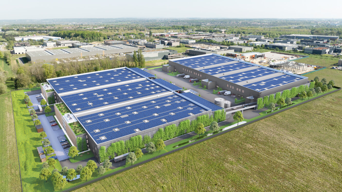 Vue aérienne d'un grand complexe industriel composé de plusieurs entrepôts gris dont les toits sont équipés de panneaux solaires, entourés de verdure et adjacents à un champ ouvert.