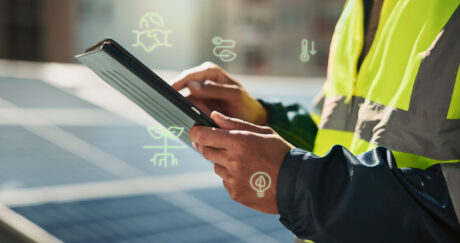 Une personne portant un gilet de haute visibilité utilise une tablette en plein air, près de panneaux solaires, avec des icônes de durabilité numérique et d'énergie superposées.