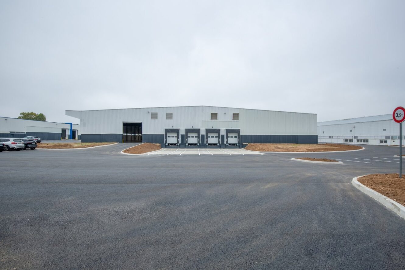 Un grand entrepôt industriel blanc avec quatre quais de chargement et un parking vide sous un ciel couvert.