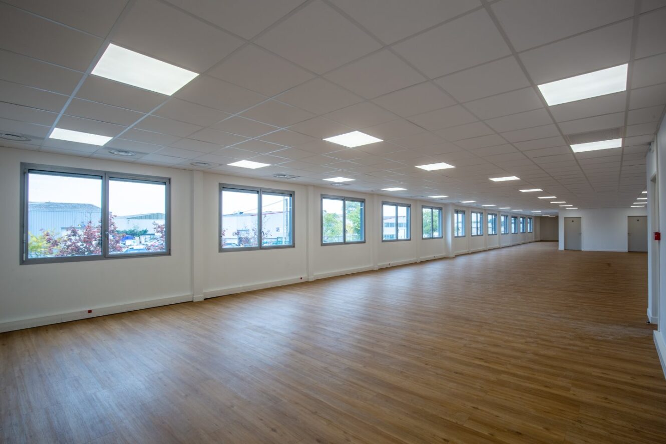 Un grand bureau vide avec un sol en bois, des murs blancs, un plafond grillagé avec des lampes fluorescentes et plusieurs grandes fenêtres sur un côté.