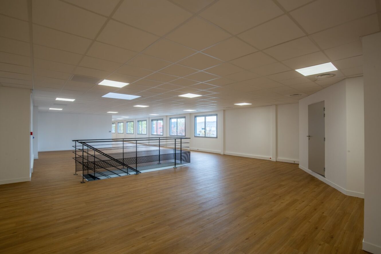 Pièce spacieuse et vide avec un sol en bois, des murs blancs, des plafonniers, plusieurs fenêtres et une rampe centrale en métal autour d'une cage d'escalier ouverte.